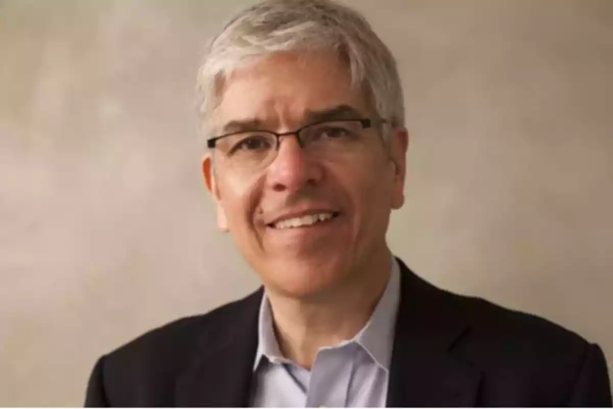 A gray-haired man with glasses smiles while wearing a dark suit and a light jersey against a neutral background. A gray-haired man with glasses smiles while wearing a dark suit and a light jersey against a neutral background.