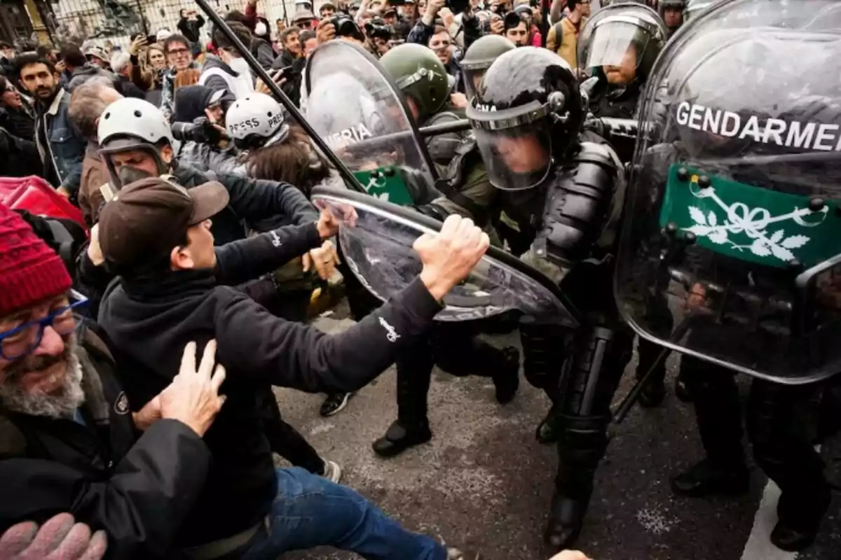 Una multitud de manifestantes se enfrenta a un grupo de gendarmes equipados con escudos y cascos en una calle concurrida. Una multitud de manifestantes se enfrenta a un grupo de gendarmes equipados con escudos y cascos en una calle concurrida.