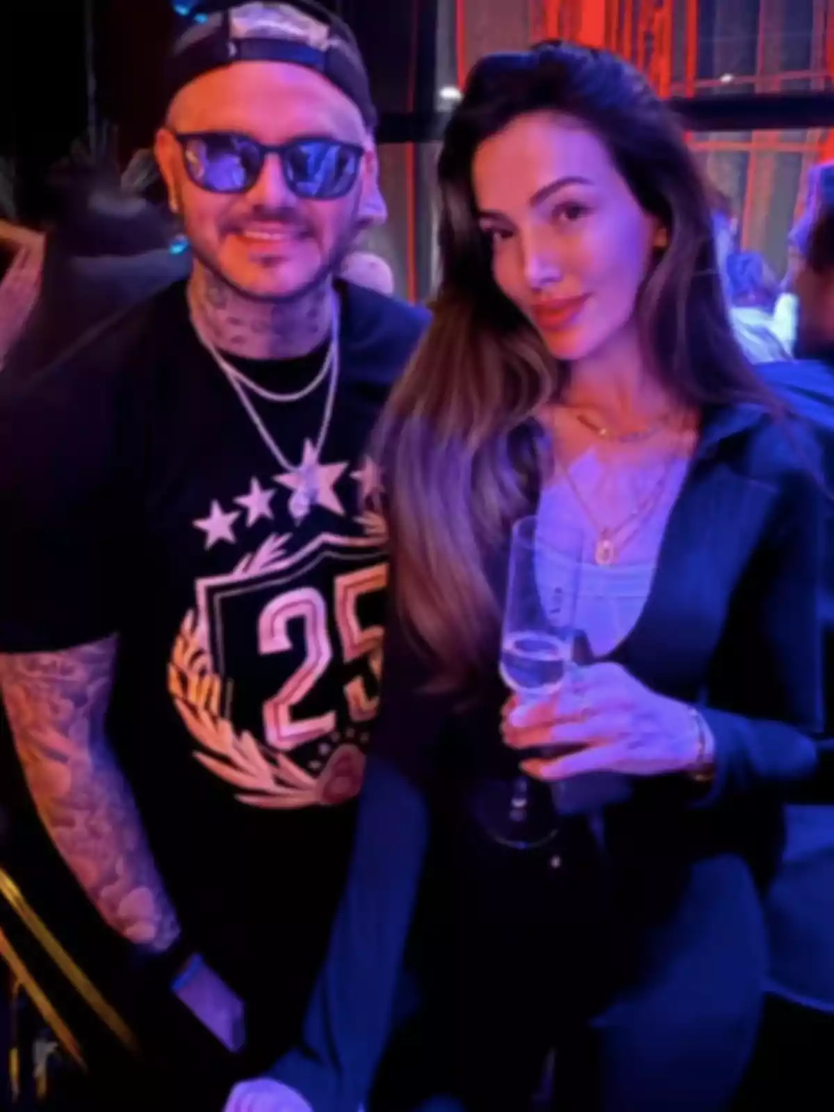 Two people pose together in a party setting, one of them wearing sunglasses and a cap, while the other holds a glass. Two people pose together in a party setting, one of them wearing sunglasses and a cap, while the other holds a glass.