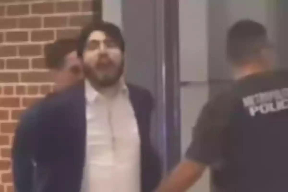 A man with a beard and glasses is being escorted by two people, one of whom is wearing a jersey with the word "POLICE," in front of a brick wall.