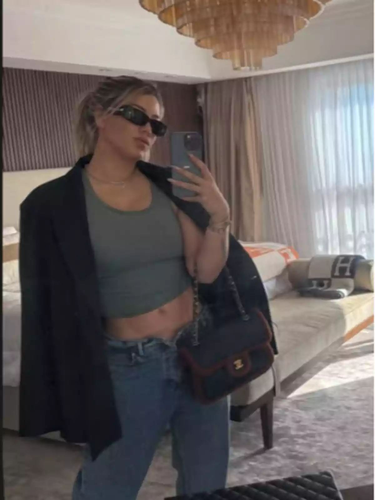 A person takes a selfie in front of a mirror in a well-lit room, wearing sunglasses, a green top, jeans, and a black blazer, while holding a handbag. A person takes a selfie in front of a mirror in a well-lit room, wearing sunglasses, a green top, jeans, and a black blazer, while holding a handbag.