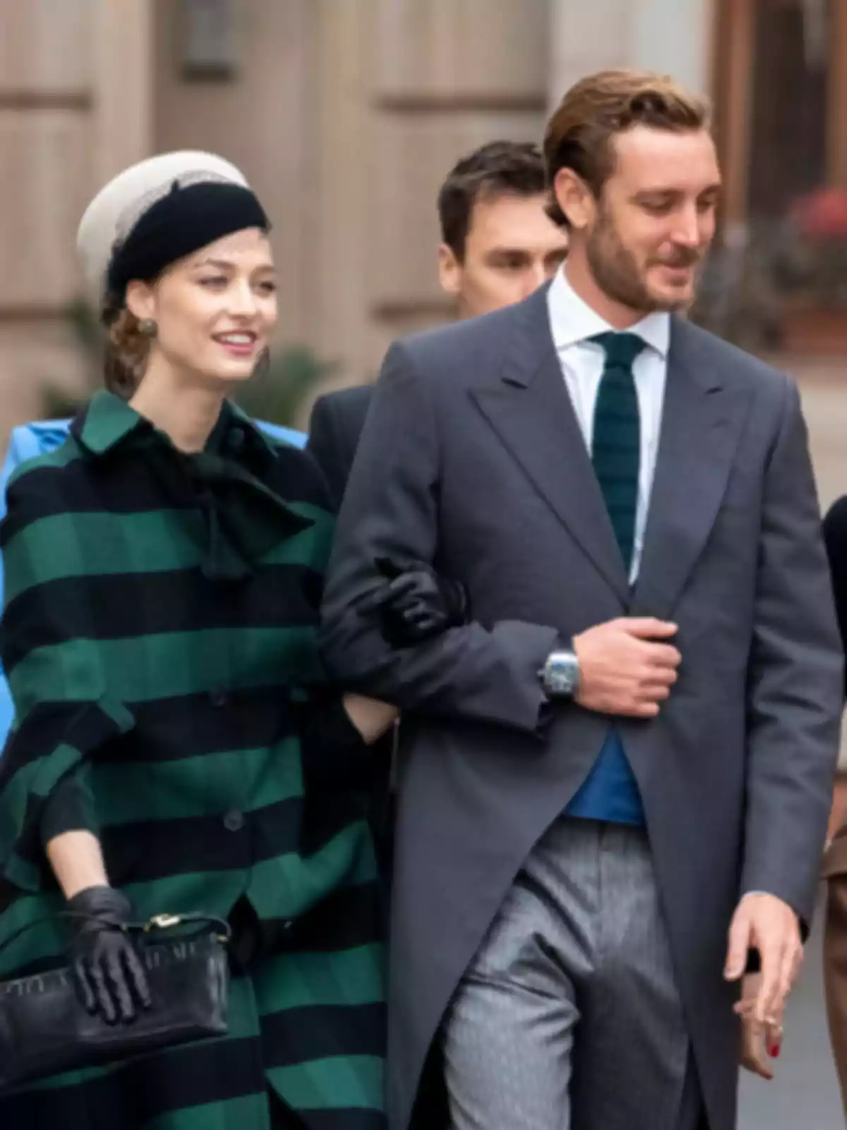 A smartly dressed couple walks down the street, she is wearing a green and black striped coat with a hat, while he is wearing a gray suit with a green tie. A smartly dressed couple walks down the street, she is wearing a green and black striped coat with a hat, while he is wearing a gray suit with a green tie.