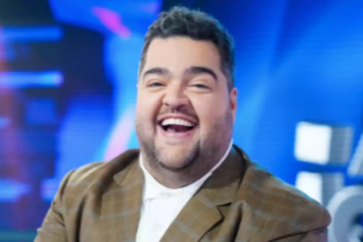A smiling man with a beard and dark hair is wearing a brown suit and a white jersey with a blurred blue background.