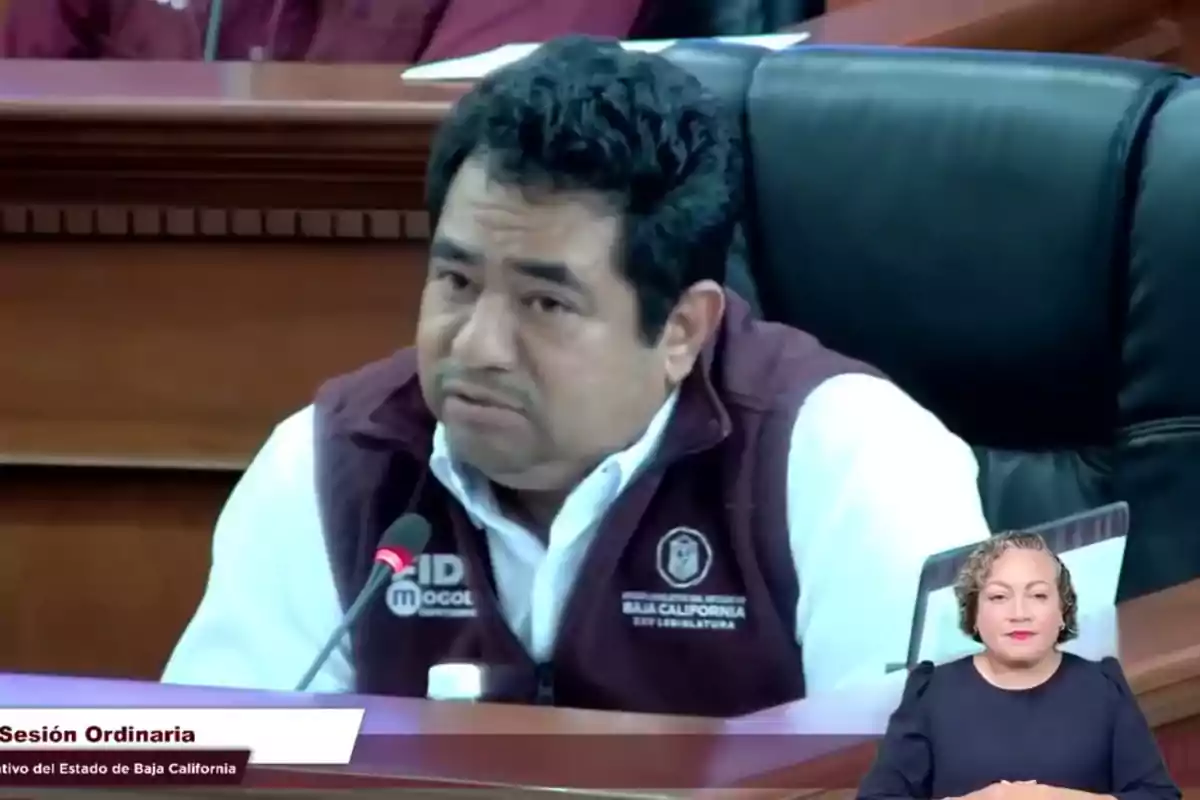 A man in a dark vest and white jersey is speaking into a microphone during a session in an official venue, while a woman in the bottom right corner is performing sign language interpretation.