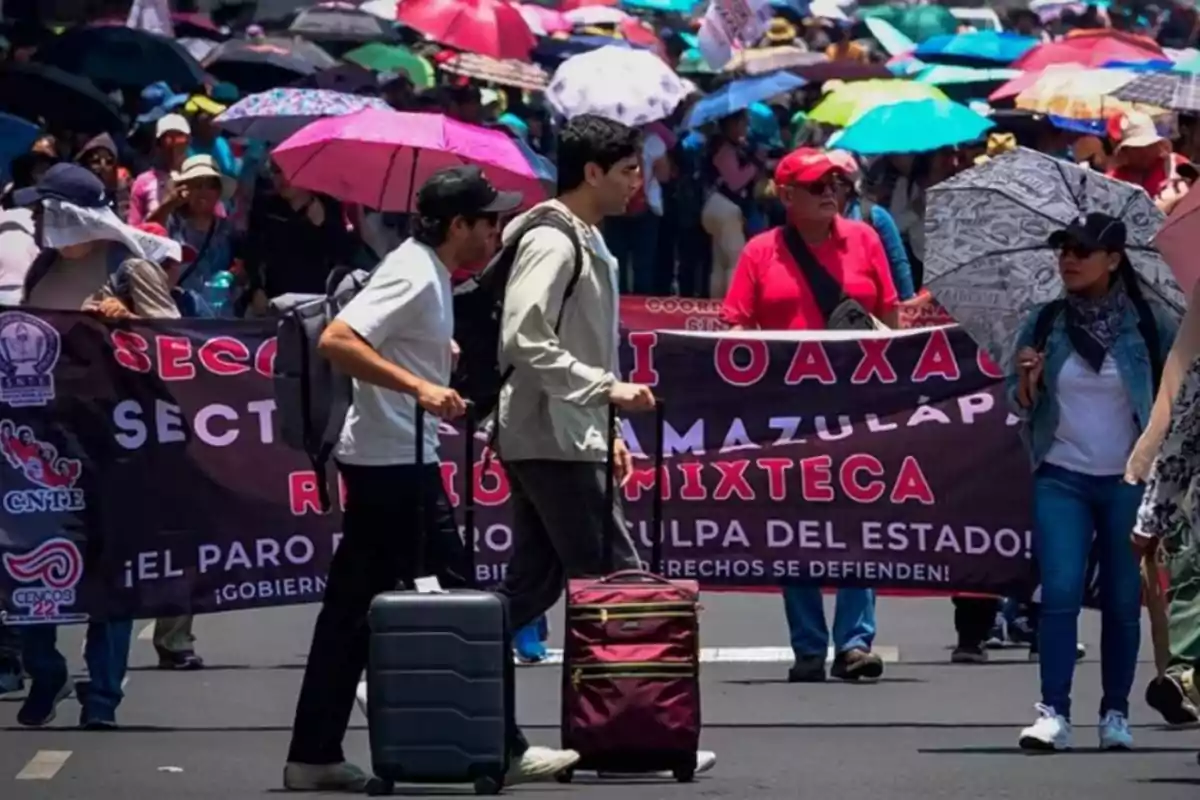 A group of people is participating in a demonstration, some are carrying colorful umbrellas and others are holding a banner with text, while two men with suitcases walk in front.