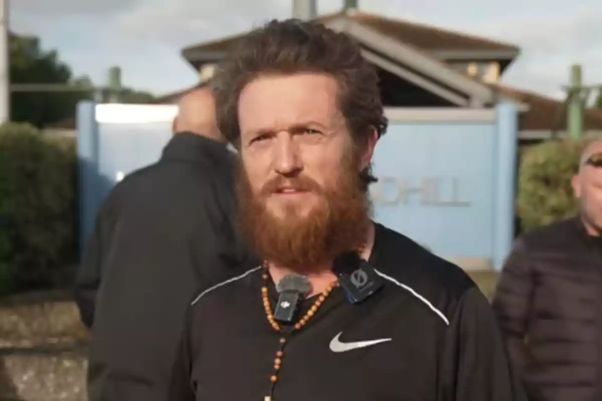 Bearded man with curly hair, wearing a black t-jersey and a necklace, standing outdoors in front of a blue building, accompanied by other people in the background.