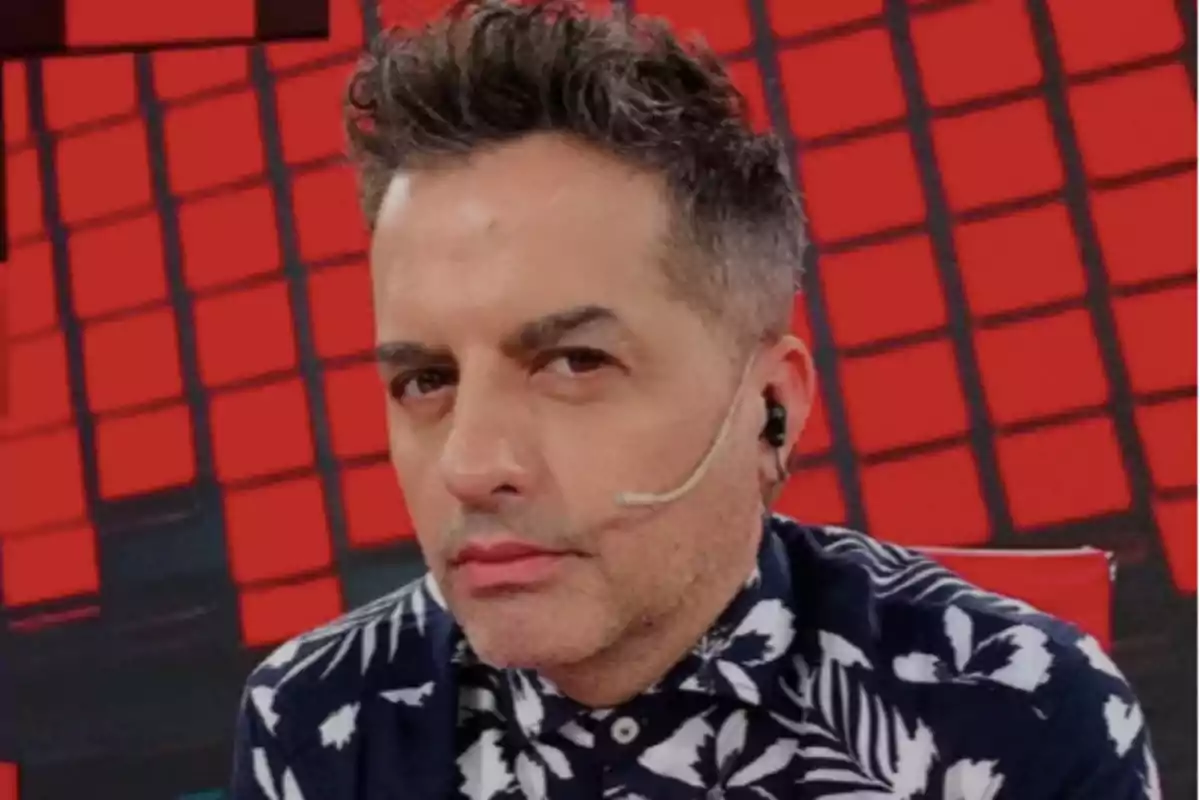 Short-haired, gray-haired man with a headset and microphone, wearing a dark jersey with a white pattern, sitting in front of a red background with a geometric pattern. Short-haired, gray-haired man with a headset and microphone, wearing a dark jersey with a white pattern, sitting in front of a red background with a geometric pattern.