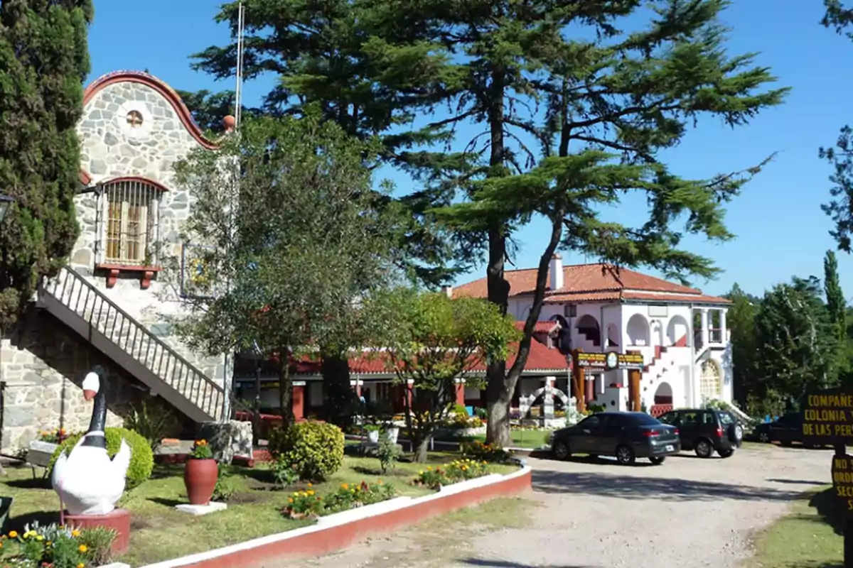 Garden with flowers and shrubs in front of a stone building and another with white walls and red roofs, surrounded by trees and parked cars Garden with flowers and shrubs in front of a stone building and another with white walls and red roofs, surrounded by trees and parked cars