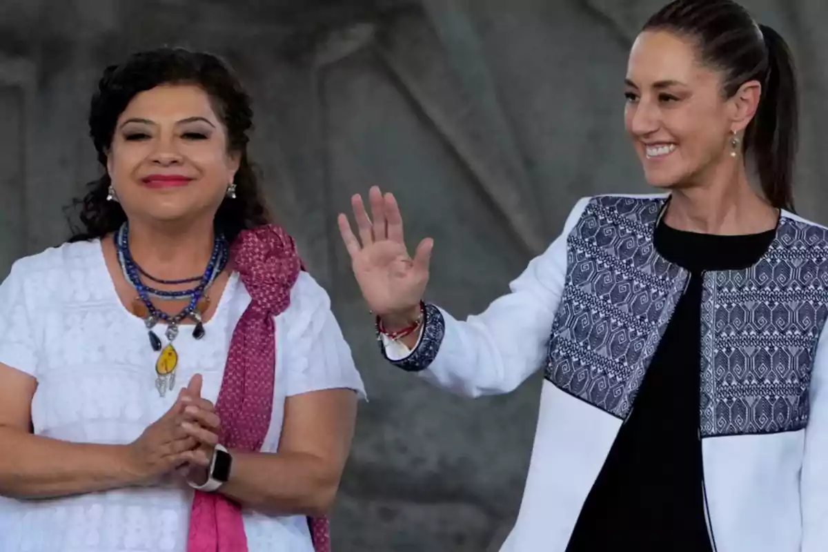 Two women smile while one of them waves, both wearing traditional and elegant clothing.