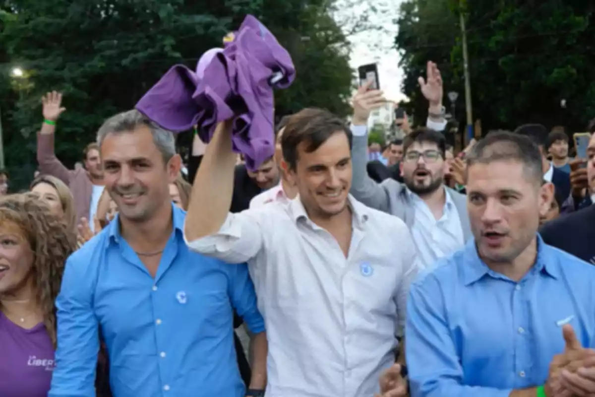 Un grupo de personas sonríe y celebra en la calle mientras un hombre en el centro levanta una prenda de color violeta Un grupo de personas sonríe y celebra en la calle mientras un hombre en el centro levanta una prenda de color violeta