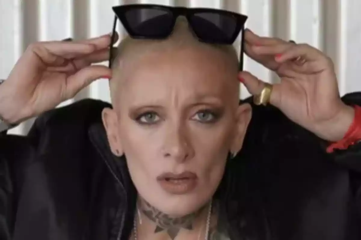 Persona de cabello rapado y ojos claros sosteniendo unas gafas de sol negras sobre su cabeza, con expresión seria y maquillaje marcado