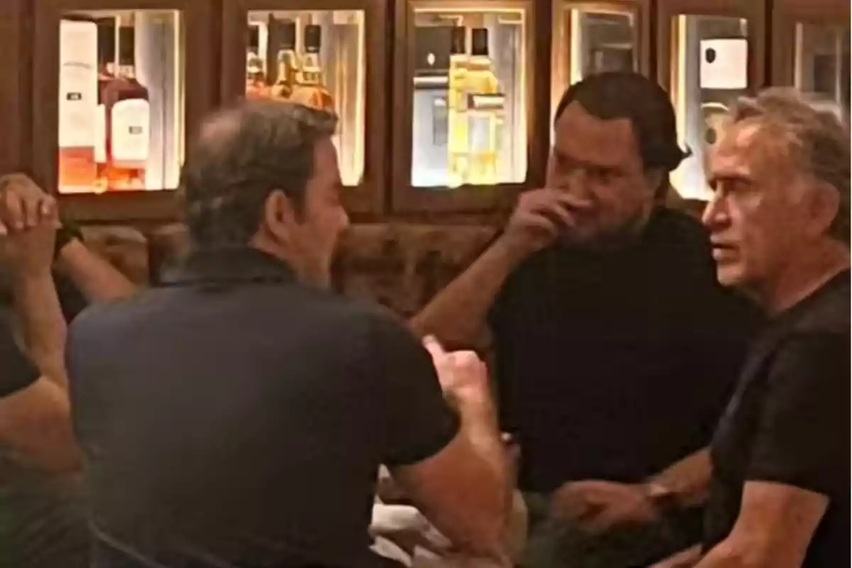 Cuatro hombres conversan sentados alrededor de una mesa en un bar con botellas iluminadas en el fondo Cuatro hombres conversan sentados alrededor de una mesa en un bar con botellas iluminadas en el fondo