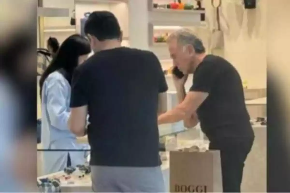 Tres personas observan objetos en una vitrina dentro de una tienda, una de ellas está hablando por teléfono y hay una bolsa de compras en primer plano
