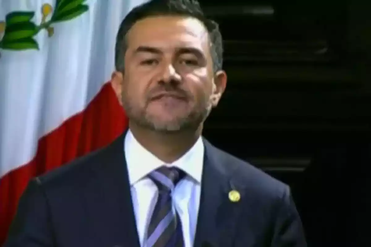 Hombre de traje y corbata hablando frente a un micrófono con una bandera de México al fondo Hombre de traje y corbata hablando frente a un micrófono con una bandera de México al fondo