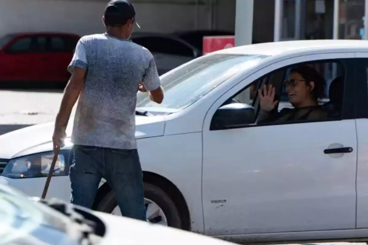 A person cleaning the windshield of a white car while the driver smiles and waves from inside the vehicle. A person cleaning the windshield of a white car while the driver smiles and waves from inside the vehicle.