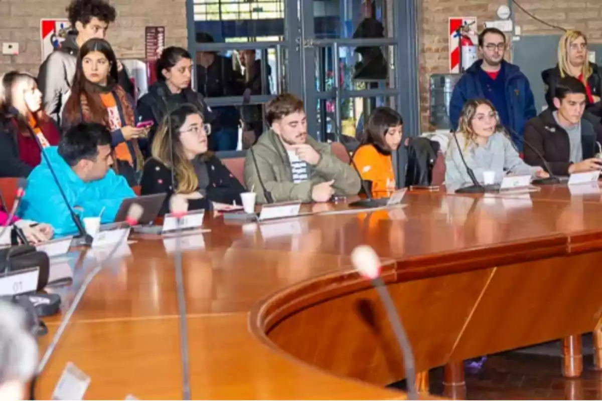 Several young people sitting around a large table in a meeting room while others are standing in the background watching.