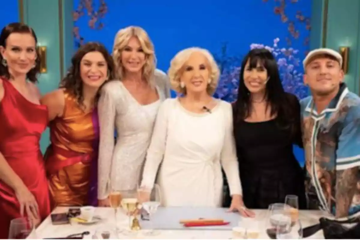 Seis personas posan sonrientes alrededor de una mesa elegantemente servida en un ambiente de estudio televisivo con fondo azul y flores. Seis personas posan sonrientes alrededor de una mesa elegantemente servida en un ambiente de estudio televisivo con fondo azul y flores.