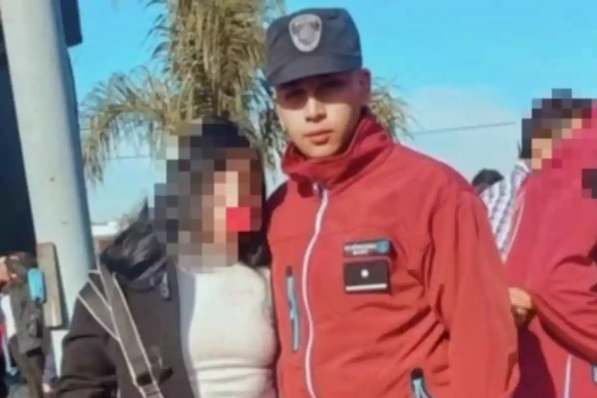 Un joven con uniforme rojo y gorra posa junto a una persona con el rostro pixelado en un entorno al aire libre con palmeras al fondo.