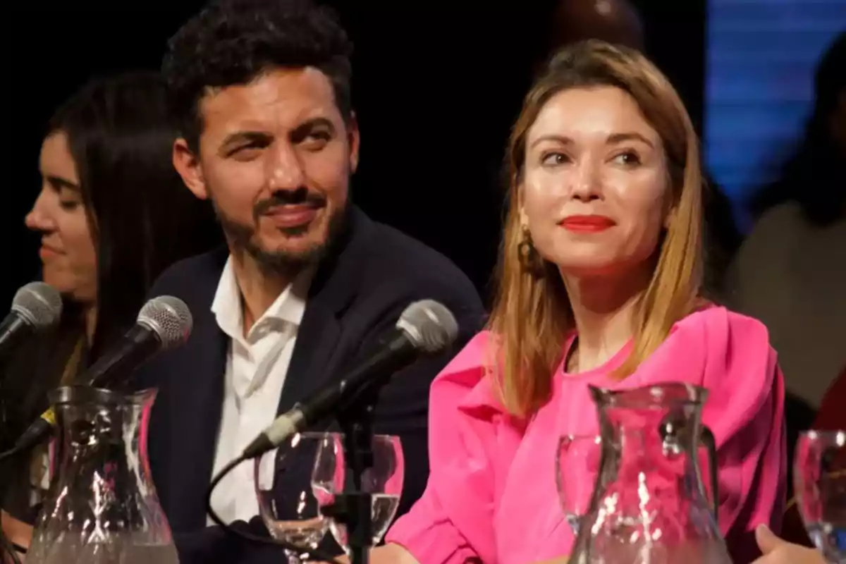 People sitting at a table with microphones and water fielders during an event or conference People sitting at a table with microphones and water fielders during an event or conference