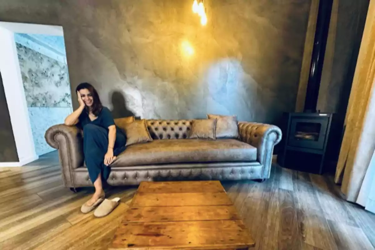 Mujer sentada en un sofá marrón en una sala con piso de madera, mesa de centro de madera y una estufa en la esquina Mujer sentada en un sofá marrón en una sala con piso de madera, mesa de centro de madera y una estufa en la esquina