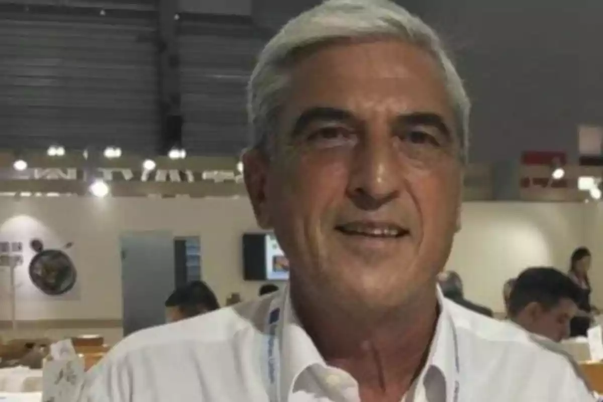 Gray-haired man in a white jersey smiling in an indoor setting with several people in the background