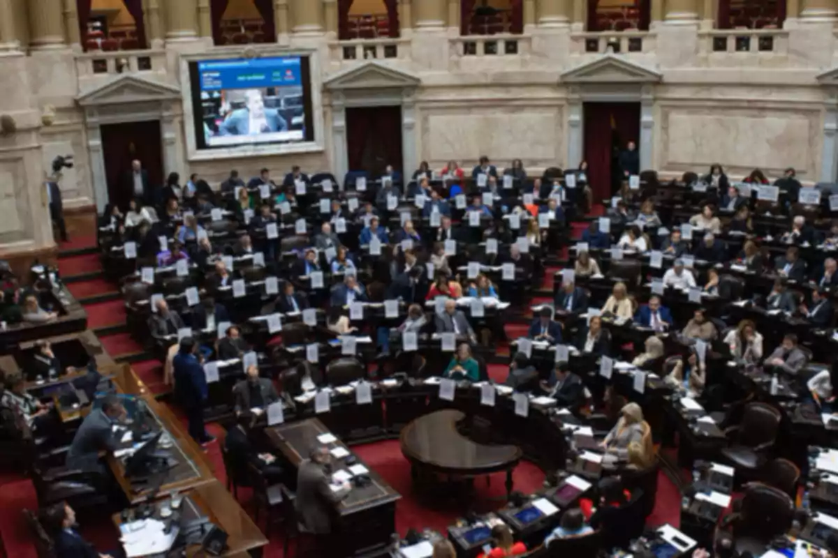Vista general de un recinto parlamentario con numerosos legisladores sentados en sus bancas y una pantalla grande al fondo mostrando una videollamada