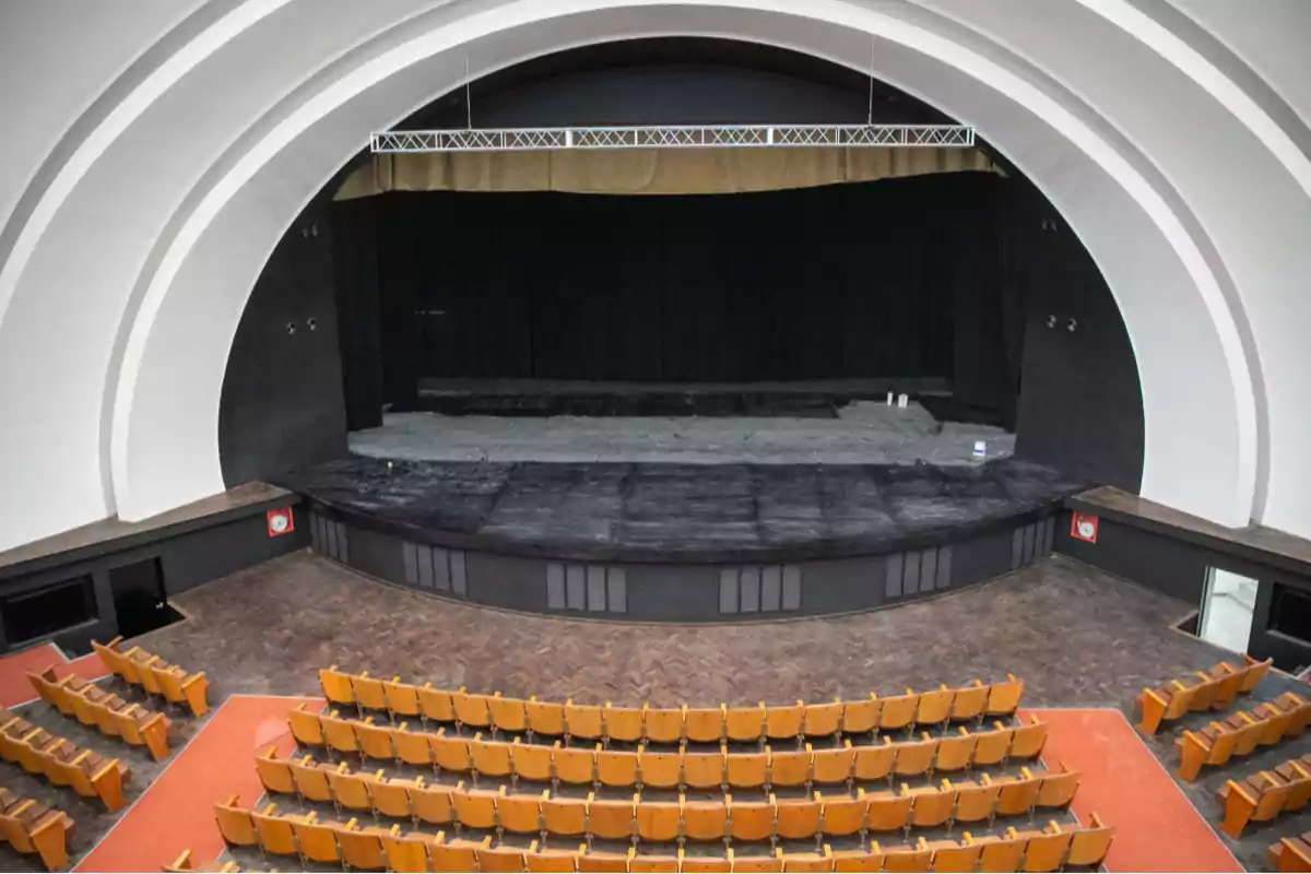 Empty auditorium with rows of wooden seats facing a large stage with black curtains and a white vaulted ceiling