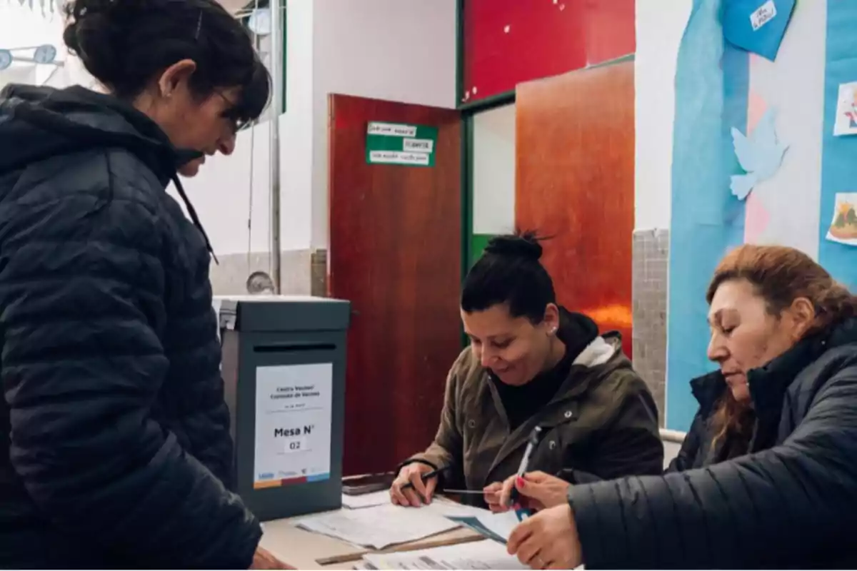 Three women are taking part in a voting process at a polling station inside a building; one of them is handing over documents while the other two are reviewing papers and lists.