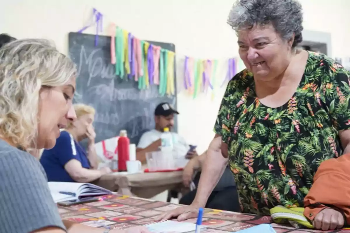 An older woman smiles while talking with another woman who is taking notes in a notebook; in the background, there are people sitting and colorful decorations on the wall.
