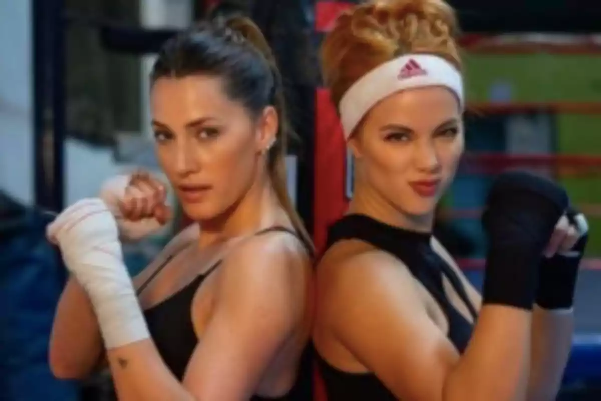 Two women in sportswear with hand wraps pose back to back in a boxing gym.