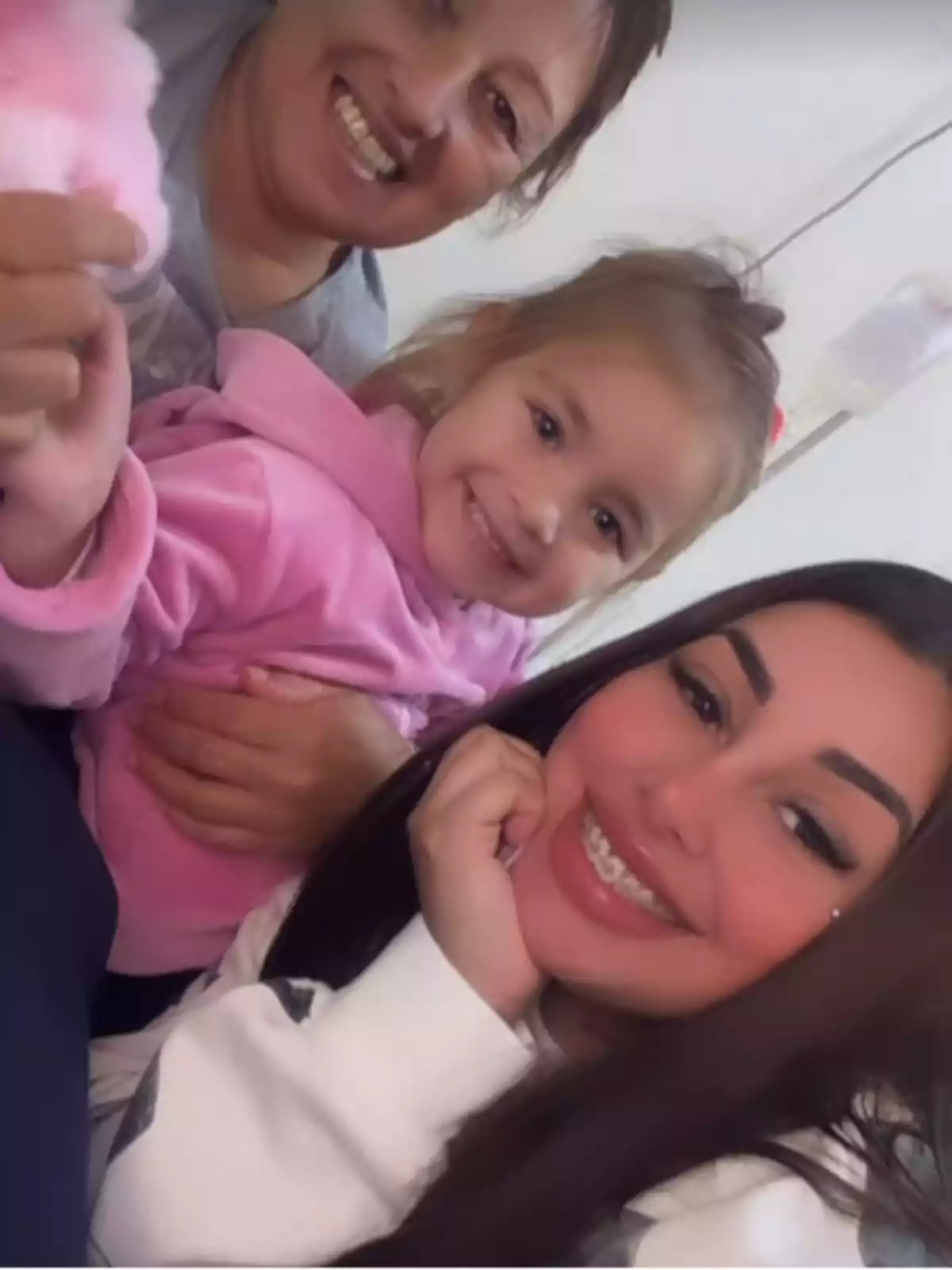 Three people smile at the camera: an older woman, a girl in a pink sweatjersey, and a young woman with braces and long dark hair.