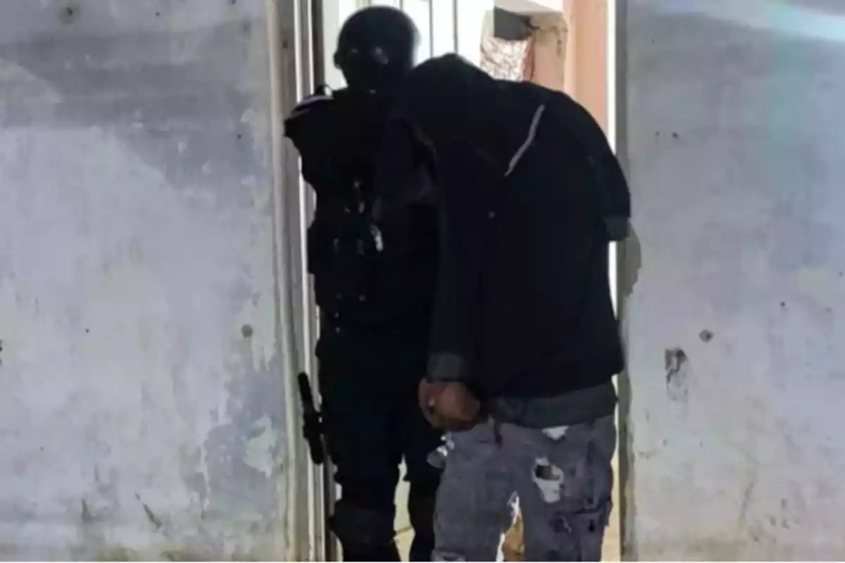 A police officer escorts a handcuffed person wearing dark clothes and torn pants inside a building with worn-out walls.