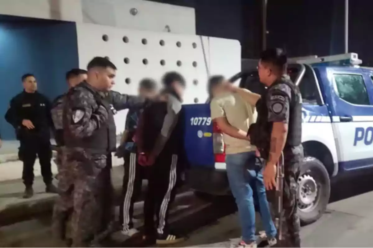 Several police officers detain three people next to a patrol car on the street at night.