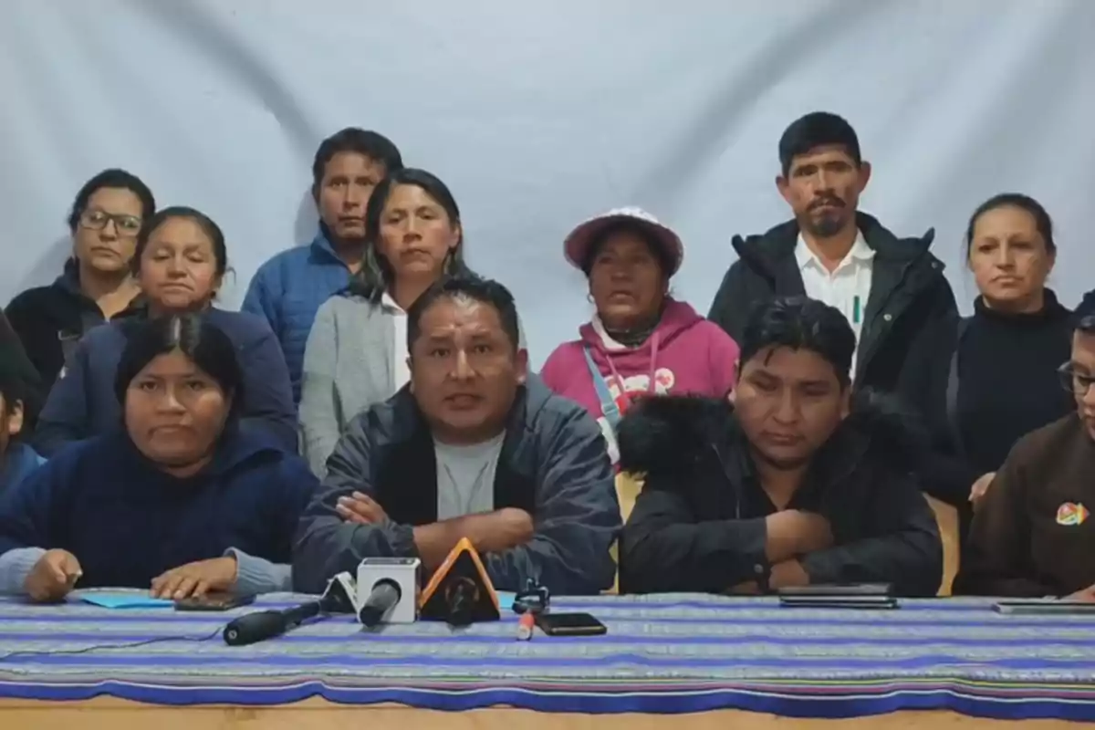 Un grupo de personas está sentado y de pie detrás de una mesa con micrófonos, mirando hacia la cámara en lo que parece ser una conferencia de prensa. Un grupo de personas está sentado y de pie detrás de una mesa con micrófonos, mirando hacia la cámara en lo que parece ser una conferencia de prensa.