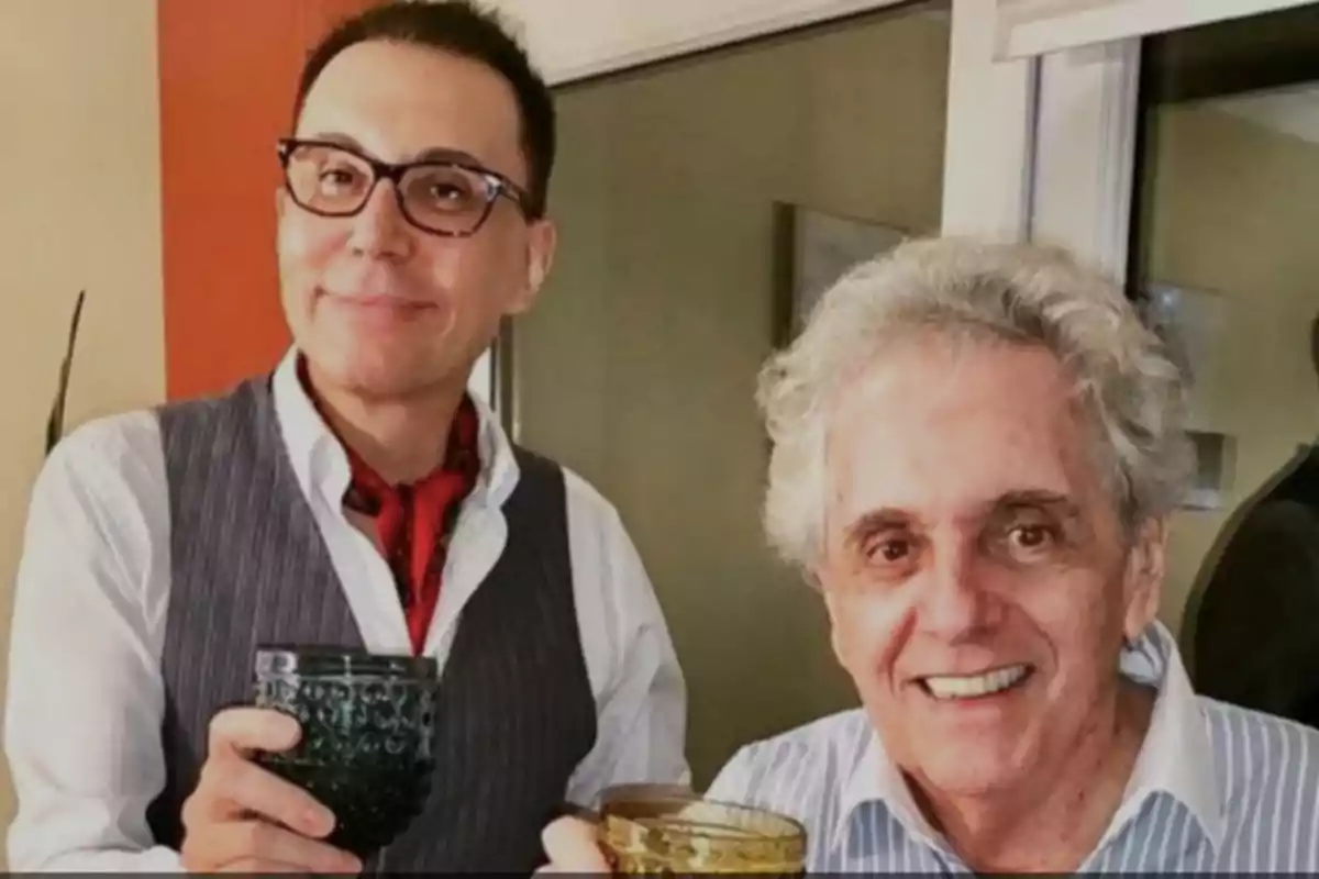 Two men smile and hold glasses at what appears to be an indoor gathering.