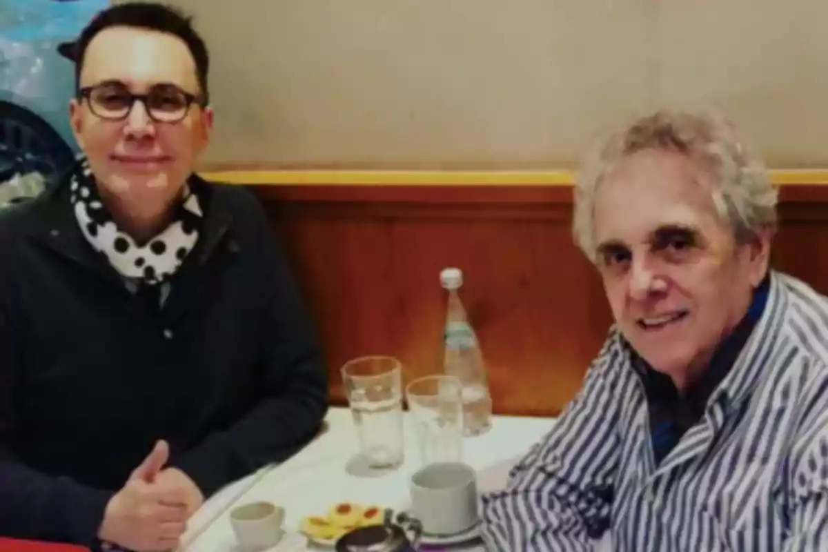 Dos hombres sentados en una mesa de restaurante con bebidas y galletas, uno de ellos lleva gafas y bufanda de lunares, el otro viste una camisa de rayas Dos hombres sentados en una mesa de restaurante con bebidas y galletas, uno de ellos lleva gafas y bufanda de lunares, el otro viste una camisa de rayas