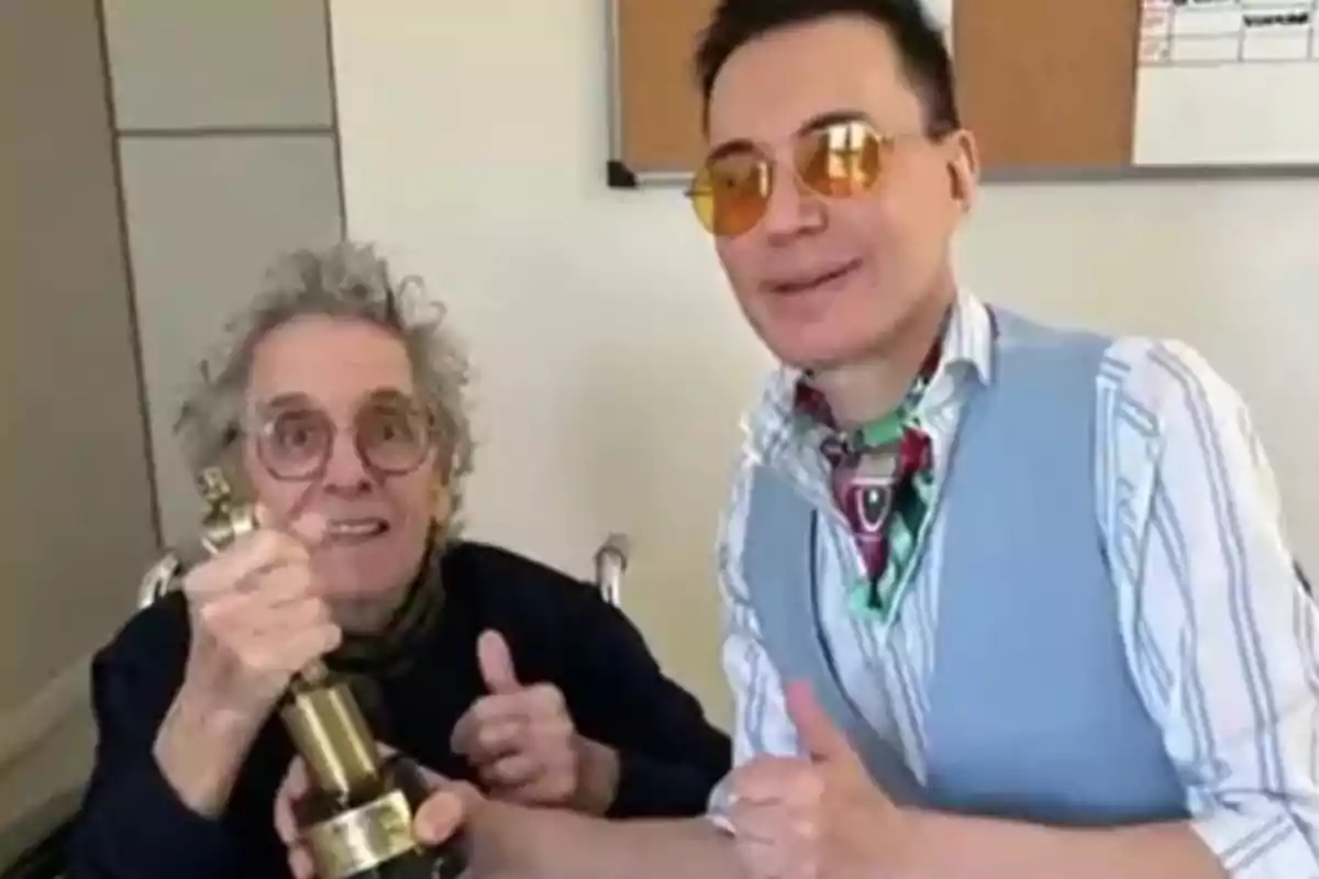 Dos personas sonríen y muestran un trofeo dorado mientras hacen señas de aprobación con el pulgar arriba en una habitación iluminada. Dos personas sonríen y muestran un trofeo dorado mientras hacen señas de aprobación con el pulgar arriba en una habitación iluminada.