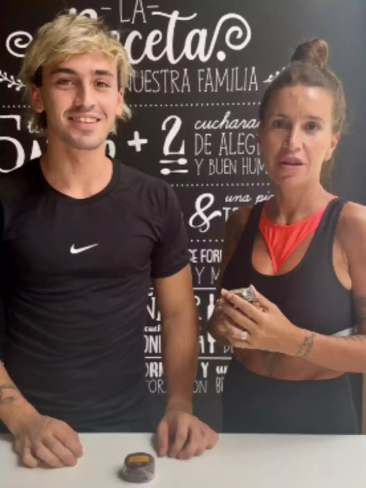 Two people pose smiling in a kitchen in front of a wall decorated with phrases and recipes; both are wearing sportswear and holding small containers on the table. Two people pose smiling in a kitchen in front of a wall decorated with phrases and recipes; both are wearing sportswear and holding small containers on the table.