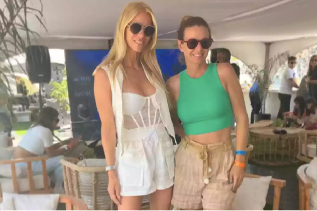 Dos mujeres posan sonrientes usando gafas de sol en un ambiente al aire libre con decoración de mimbre y otras personas al fondo Dos mujeres posan sonrientes usando gafas de sol en un ambiente al aire libre con decoración de mimbre y otras personas al fondo