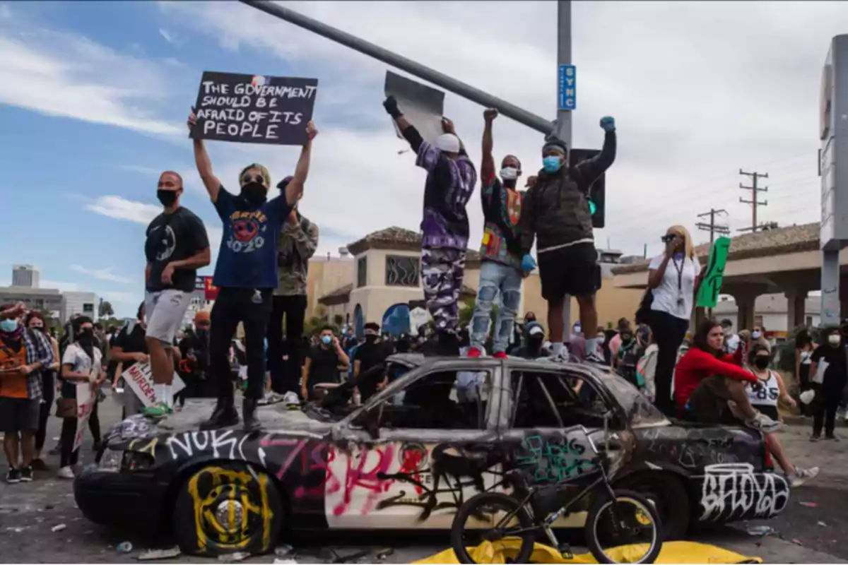 Personas manifestándose y posando sobre un coche quemado y cubierto de grafitis en una calle urbana, algunas sostienen pancartas y la mayoría lleva mascarillas. Personas manifestándose y posando sobre un coche quemado y cubierto de grafitis en una calle urbana, algunas sostienen pancartas y la mayoría lleva mascarillas.