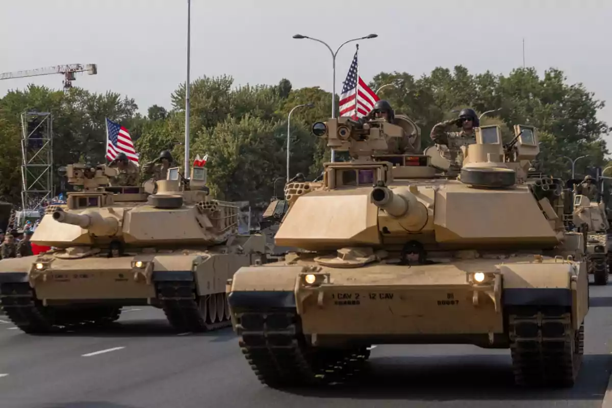 U.S. military tanks move down an avenue during a parade with soldiers on board and American flags waving.