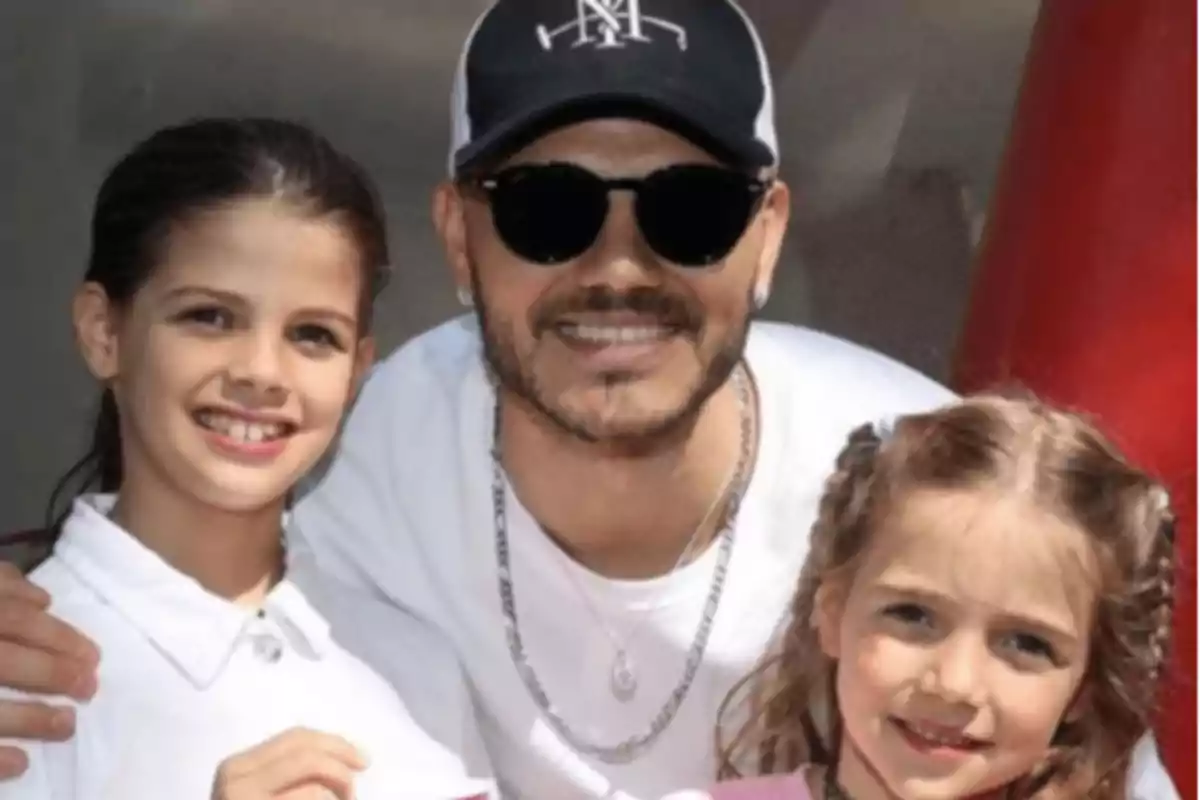 Hombre con gafas de sol y gorra negra sonríe junto a dos niñas pequeñas que también están sonriendo Hombre con gafas de sol y gorra negra sonríe junto a dos niñas pequeñas que también están sonriendo