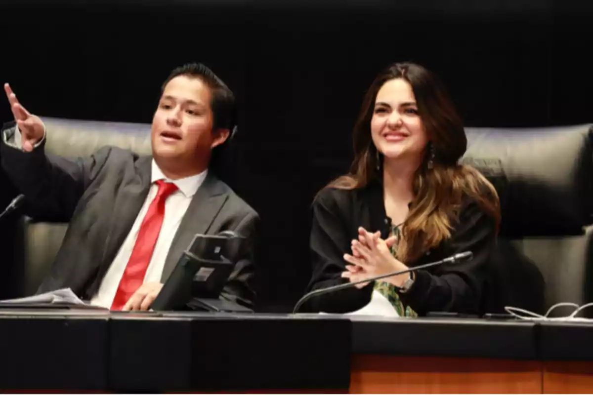 Dos personas sentadas en lo que parece ser un recinto legislativo, una de ellas viste traje y corbata roja y está señalando hacia el frente mientras la otra persona sonríe y aplaude Dos personas sentadas en lo que parece ser un recinto legislativo, una de ellas viste traje y corbata roja y está señalando hacia el frente mientras la otra persona sonríe y aplaude