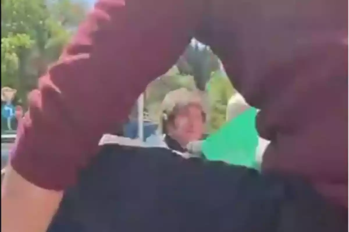 Persona en el fondo mirando hacia la cámara mientras otra persona con suéter rojo aparece en primer plano en un entorno al aire libre con árboles Persona en el fondo mirando hacia la cámara mientras otra persona con suéter rojo aparece en primer plano en un entorno al aire libre con árboles