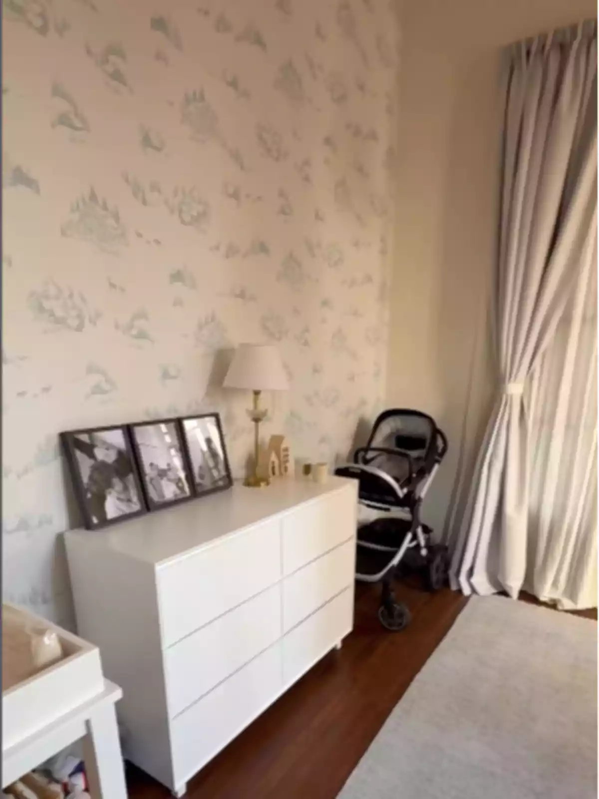 Habitación infantil decorada con papel tapiz claro, cómoda blanca con fotos enmarcadas y una carriola junto a una ventana con cortinas largas Habitación infantil decorada con papel tapiz claro, cómoda blanca con fotos enmarcadas y una carriola junto a una ventana con cortinas largas