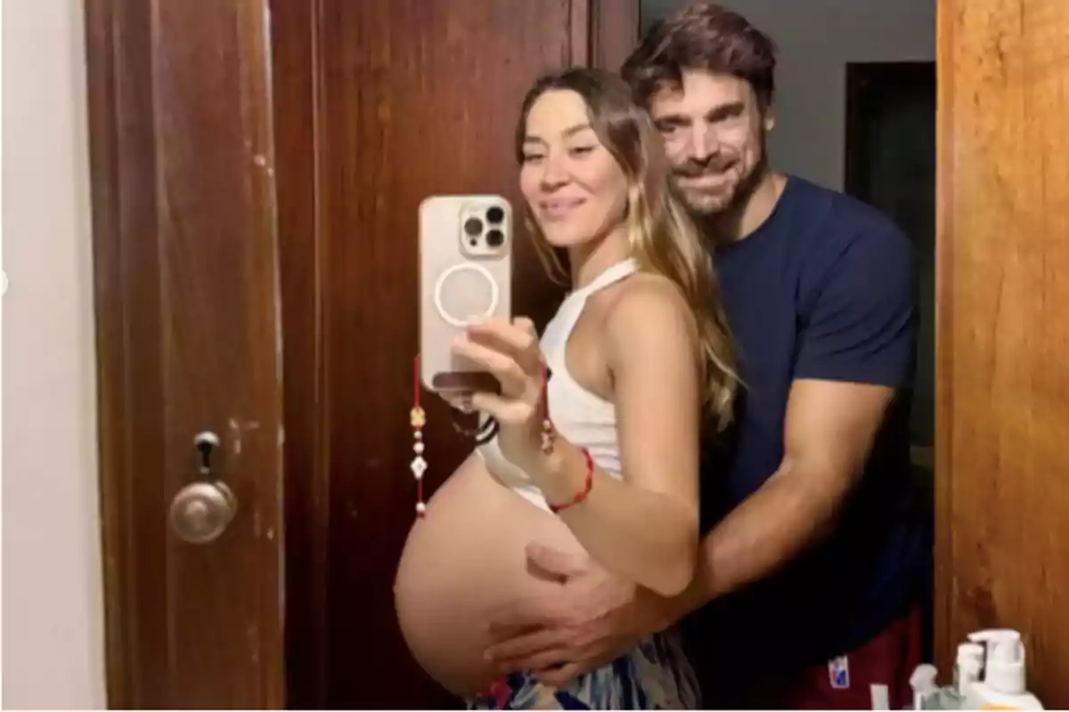 Pareja sonriente tomándose una selfie en el espejo mientras la mujer embarazada muestra su vientre y el hombre la abraza por detrás Pareja sonriente tomándose una selfie en el espejo mientras la mujer embarazada muestra su vientre y el hombre la abraza por detrás