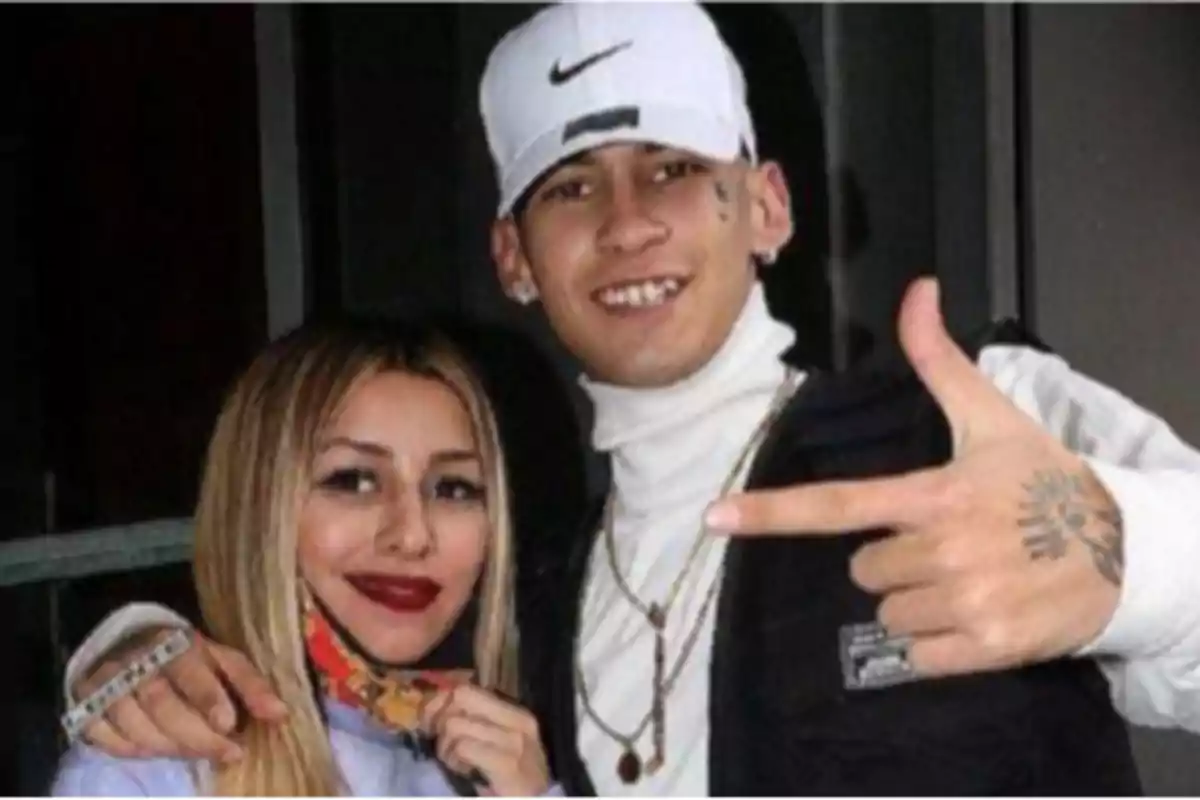 A woman and a man pose together smiling; the man is wearing a white cap and making a gesture with his hand, while the woman is holding a face mask below her face.