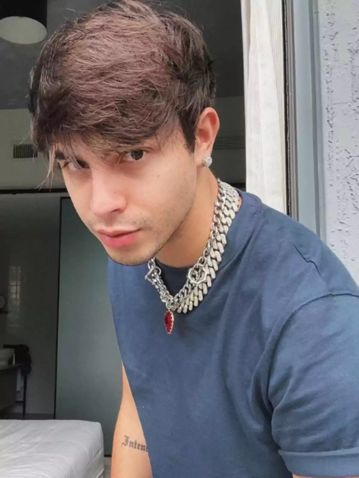 Joven con cabello castaño y camiseta azul posando cerca de una ventana, lleva un collar grueso plateado y un pendiente brillante en la oreja izquierda Joven con cabello castaño y camiseta azul posando cerca de una ventana, lleva un collar grueso plateado y un pendiente brillante en la oreja izquierda