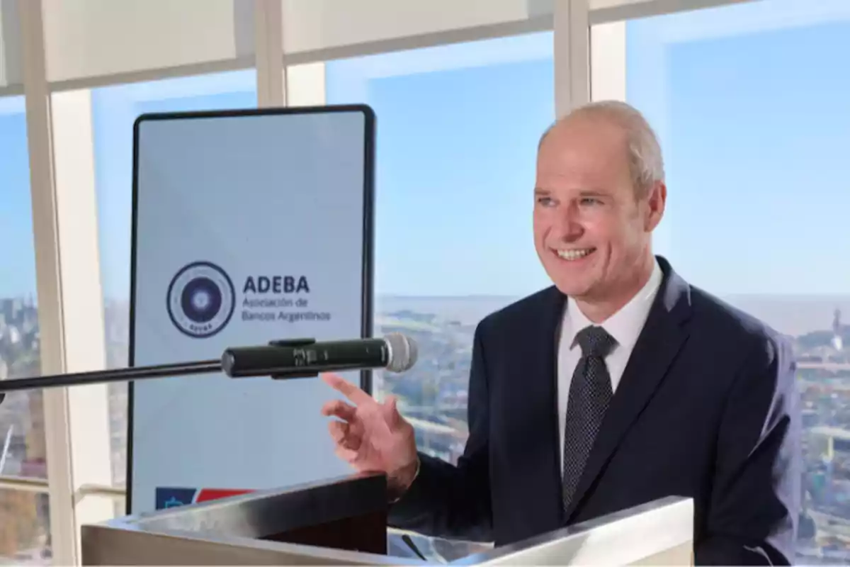 Man in a suit smiling while giving a speech at a podium with a microphone and a screen in the background displaying the logo of the Association of Argentine Banks in a room with large windows and a view of the city Man in a suit smiling while giving a speech at a podium with a microphone and a screen in the background displaying the logo of the Association of Argentine Banks in a room with large windows and a view of the city
