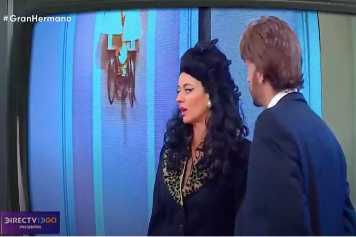 Two elegantly dressed people are talking in front of a blue wall while the show Big Brother is being broadcast on television. Two elegantly dressed people are talking in front of a blue wall while the show Big Brother is being broadcast on television.